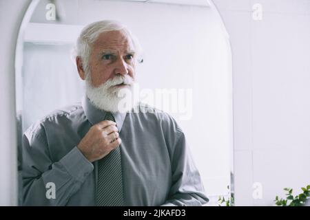 Ritratto di bell'uomo anziano con barba bianca guardando lo specchio mentre si regola la cravatta Foto Stock
