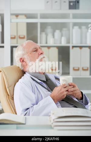 Medico anziano stanco che si rilassa sopra una tazza di tè nel suo ufficio Foto Stock