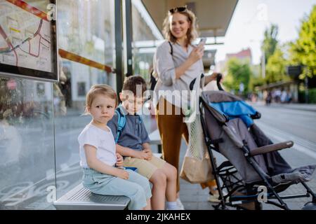 Giovane madre con bambini in attesa sulla fermata dell'autobus in città. Foto Stock