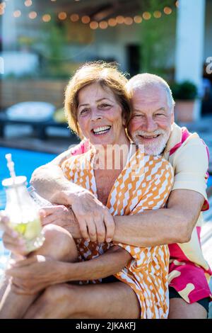 Coppia anziana felice di godere di bevande quando si rilassa e seduta in piscina in estate. Foto Stock