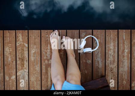 Primo piano delle gambe e delle cuffie del woman che giacciono su un molo vicino al lago naturale in estate vista dall'alto. Foto Stock