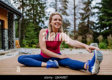 Giovane donna che pratica yoga, che si estende su un patio cottage di montagna circondato dalla natura. Foto Stock
