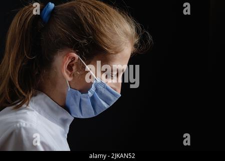 Un medico donna in una maschera protettiva è sconvolto o piangere. Un infermiere in una maschera viene premuto su uno sfondo nero. Medico stanco di mezza età su ba scuro Foto Stock