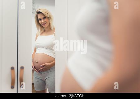 donna incinta felice guardando nello specchio a casa, medio full shot gravidanza concetto. Foto di alta qualità Foto Stock