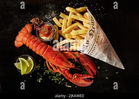 Vista dall'alto dell'aragosta rossa yummy e patatine fritte in foglio di giornale servito con salsa ed erbe vicino alle fette di lime su sfondo nero Foto Stock