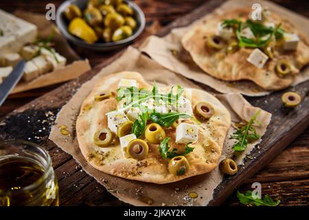 Alto angolo di pane piatto appena sfornato con formaggio feta stagionato pezzi di olive e rucola servito sul tagliere vicino ingrediente in cucina Foto Stock