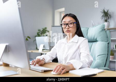Apprendimento a distanza online. Giovane bella insegnante femminile asiatica in cuffie e occhiali seduti a un tavolo ad un computer che insegna una lezione di classe online. Foto Stock