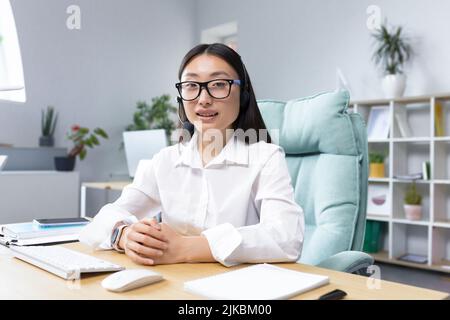 Ritratto di una giovane donna asiatica bella in occhiali e cuffie con microfono, seduto in ufficio alla scrivania conduzione di un webinar online, consulenza, raccontare, gesturing con le mani. Foto Stock
