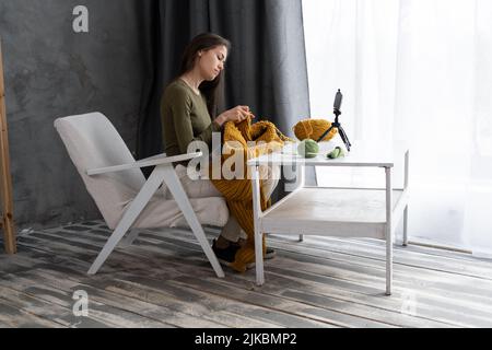 blogger ragazza registra video di maglieria utilizzando smartphone su treppiede, la donna impara a lavorare a maglia visitando classe online, blog e hobby Foto Stock