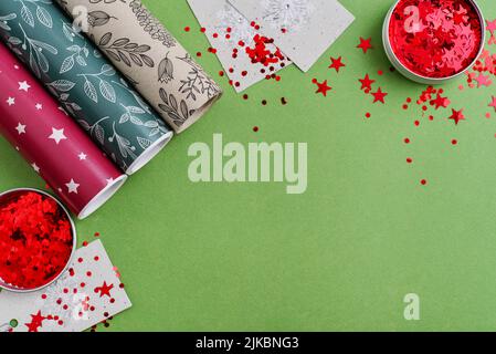 Rotoli di carta da imballaggio colorata con cartellini regalo e confetti rossi scintillanti su sfondo verde, vista dall'alto Foto Stock