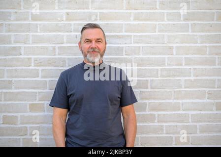 Ritratto della parte superiore del corpo di un uomo anziano con bearded in camicia estiva blu che si posa davanti ad un muro di mattoni Foto Stock