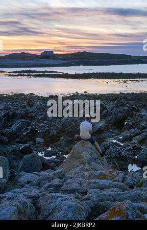 Un uomo che guarda il sole tramontare sull'isola di Lihou da l'Erée, Guernsey, Isole del canale Foto Stock