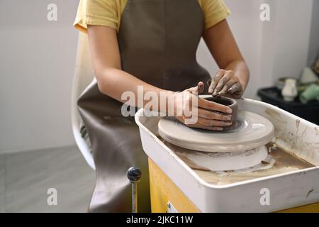 Vista ritagliata della giovane donna che indossa un grembiule che crea una ciotola di ceramica fatta a mano in un laboratorio di ceramica Foto Stock