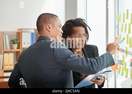 Gruppo di lavoratori concentrati di colletti bianchi in piedi su tavola di vetro e leggere note appiccicose mentre si ha una riunione di lavoro in grande spazio sala riunioni Foto Stock