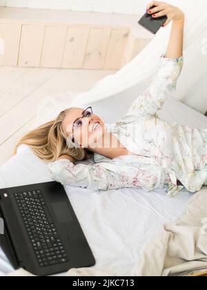 Giovane donna beauty blogger prende un selfie mentre si trova a letto. Si è svegliata e chiacchiera con i suoi seguaci durante lo streaming in linea. Foto verticale. Foto Stock