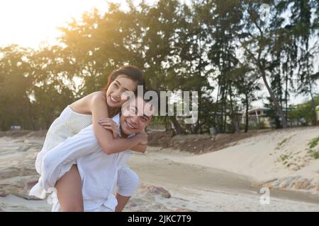 Godendo la vacanza in un resort esotico: Bell'uomo asiatico con ampio sorriso guardando via mentre dando giro piggyback alla sua bella ragazza Foto Stock