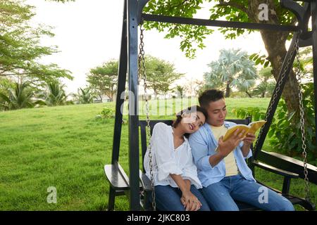 Adorabile coppia asiatica indossando jeans e camicie seduti sul portico swing e godendosi a vicenda compagnia, bel giovane uomo avvolto in libro lettura, verde parco pubblico sullo sfondo Foto Stock
