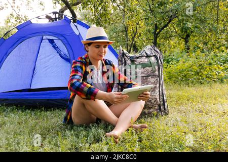 Defocus giovane donna che lavora sul tablet vicino campeggio tenda all'aperto circondato da bella natura. Freelance, sabbatico, salute mentale. OK gesto. R Foto Stock