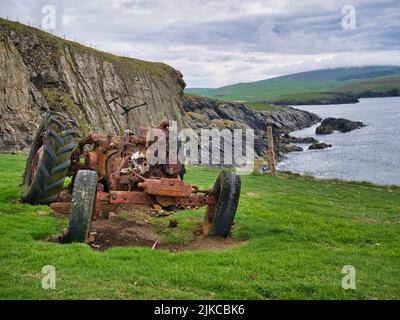I resti di un trattore abbandonato e arrugginito in un campo erboso sulla costa vicino a Bigton, Shetland, Regno Unito. Preso in una giornata nuvolosa e colma. Foto Stock