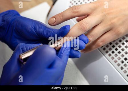 Il manicurista dipinge le unghie beige con vernice ibrida in guanti di lattice blu. Foto Stock