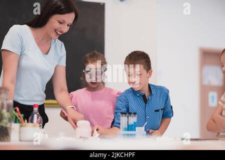 Scuola elementare Scienza Aula: Insegnante entusiasta spiega la chimica a diversi gruppi di bambini, Little Boy Mixes Chemicals in Beakers Foto Stock