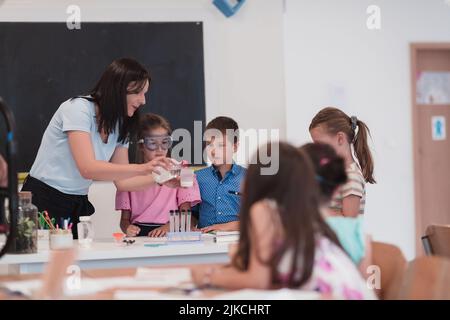 Scuola elementare Scienza Aula: Insegnante entusiasta spiega la chimica a diversi gruppi di bambini, Little Boy Mixes Chemicals in Beakers Foto Stock