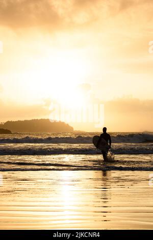 Surfista camminando sulla spiaggia al tramonto a Tofino, British Columbia Foto Stock