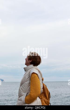 Donna matura felice in abiti gialli sta in piedi vicino al mare Foto Stock