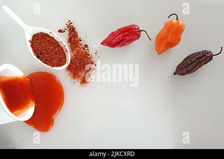 Tre varietà di peperoni caldi Scotch cofano, naga morich, bhut jolokiaand loro derivati - salsa e pepe caldo macinato su una superficie di vetro bianco Foto Stock