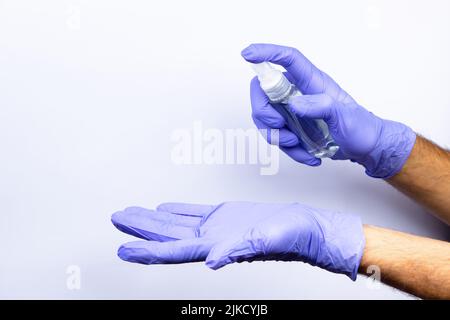 La mano di un uomo in un guanto medico blu spruzza un antisettico da una fiala su una seconda mano guantata. Su sfondo bianco. Un sacco di spazio vuoto Foto Stock