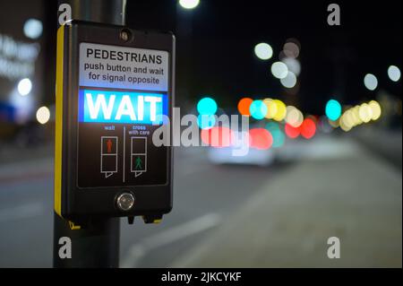 Pulsante di attraversamento pedonale e cartello di attesa illuminato con traffico fuori fuoco sullo sfondo Foto Stock