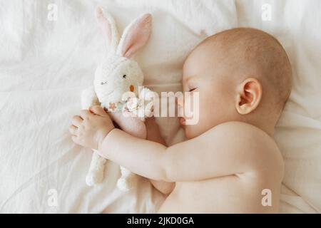 Bimbo addormentato con il giocattolo morbido preferito a portata di mano. Sonno spensierato del bambino con il giocattolo morbido sul letto. Bambino che dorme su un lato nel letto a casa, nuova famiglia Foto Stock