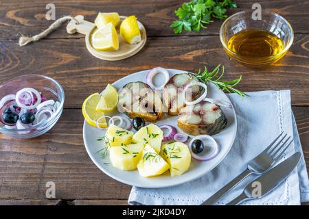 Sgombro affumicato tagliato su piatto con patate bollite, anelli di cipolla, limone, olive, olio, verdi su sfondo di legno Foto Stock