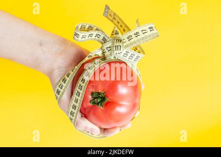 mano della donna che tiene un pomodoro, legato con un arco di metro a nastro, isolato su sfondo giallo. Foto Stock