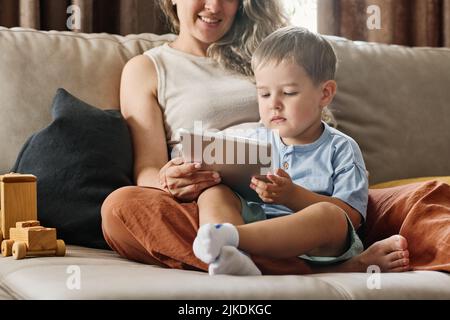 Serio bambino in casualwear guardando lo schermo di tavoletta tenuto da sua madre mentre sia seduto sul divano e guardare cartoni animati Foto Stock