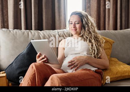 Felice giovane donna incinta che guarda lo schermo del tablet durante le comunicazioni video con i suoi colleghi o amici a casa Foto Stock