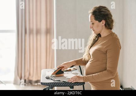 Vista laterale della giovane donna incinta in abito casual che stira il baby pullover mentre si trova di fronte all'asse in casa Foto Stock