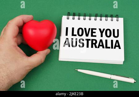 Sulla superficie verde ci sono una penna, un blocco note con l'iscrizione - ti amo Australia. Un uomo ha un cuore nelle sue mani. Foto Stock