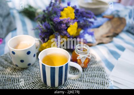 bouquet estivo di yarrow giallo e lavanda viola, 2 tazze a righe e pallini di tè con latte e vasetto di marmellata di mandarino su una coperta da picnic a righe con atmosfera estiva Foto Stock