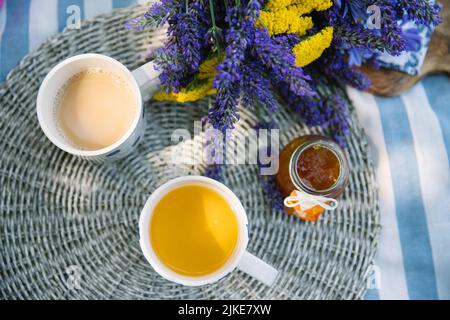 bouquet estivo di yarrow giallo e lavanda viola, 2 tazze di tè con latte e vasetto di marmellata di mandarino su una coperta da picnic a strisce con atmosfera estiva Foto Stock