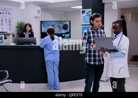 Medico afroamericano che tiene il laptop che presenta i risultati di laboratorio al paziente asiatico in clinica privata di pratica. Medico che parla con il giovane uomo che controlla l'appuntamento sul computer portatile. Foto Stock