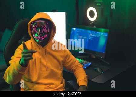 Giovane uomo nero con cappuccio arancione e strana maschera che indica la fotocamera seduta dalla scrivania con impostazione di gioco e la ghiera di luce. Illuminazione verde scuro. Foto di alta qualità Foto Stock