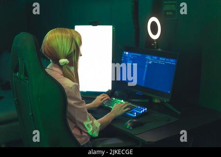 Ragazza bianca adolescente che indossa graziosi ponytail seduti in sedia da gioco utilizzando un computer professionale con luce ad anello. Illuminazione verde scuro. Foto di alta qualità Foto Stock