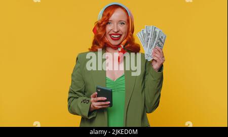 Felice redhead giovane donna guardando smartphone mostra sinceramente gioire vincere, ricevere denaro in denaro banconote in denaro, successo lotteria fortuna. Zenzero ragazza studio interno isolato su sfondo giallo parete Foto Stock