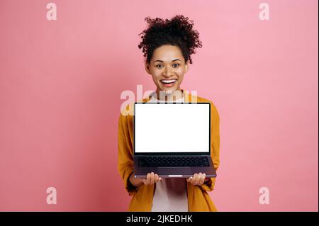 Stupito felice afroamericana giovane donna in elegante vestito, tenendo aperto il laptop con schermo bianco vuoto, si leva su isolato sfondo rosa, guarda la fotocamera, mockup per la tua pubblicità, spazio copia Foto Stock