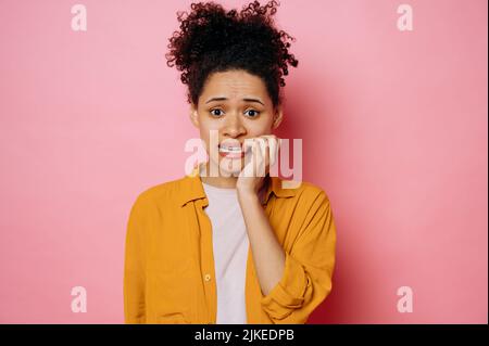 Confuso preoccupato afroamericana giovane donna, in abbigliamento casual, guardando stressato e nervoso con le mani sulla bocca mordente unghie, guardando la macchina fotografica, sta attraversando, si erge su isolato sfondo rosa Foto Stock