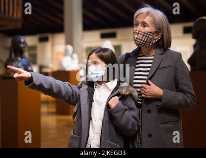 Tween studentessa e anziana tutor femminile in maschere facciali osservando la mostra in galleria d'arte Foto Stock