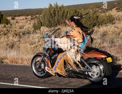 L'uomo nativo americano vestito tradizionalmente (Arapahoe) guida la motocicletta Harley Davidson lungo l'autostrada di campagna Foto Stock