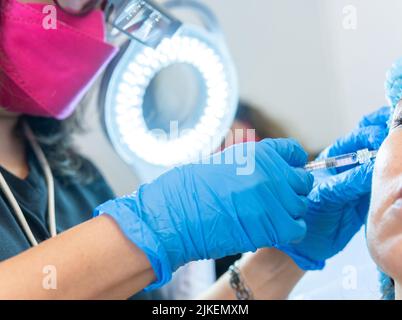 Primo piano di una dottoressa che applica acido ialuronico con una siringa ad un paziente illuminato da una lampada a LED per dermatologia Foto Stock