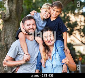 Il bambino che vive con sicurezza, impara ad avere fede. Ritratto di una bella famiglia con i loro figli sulle spalle in un parco. Foto Stock